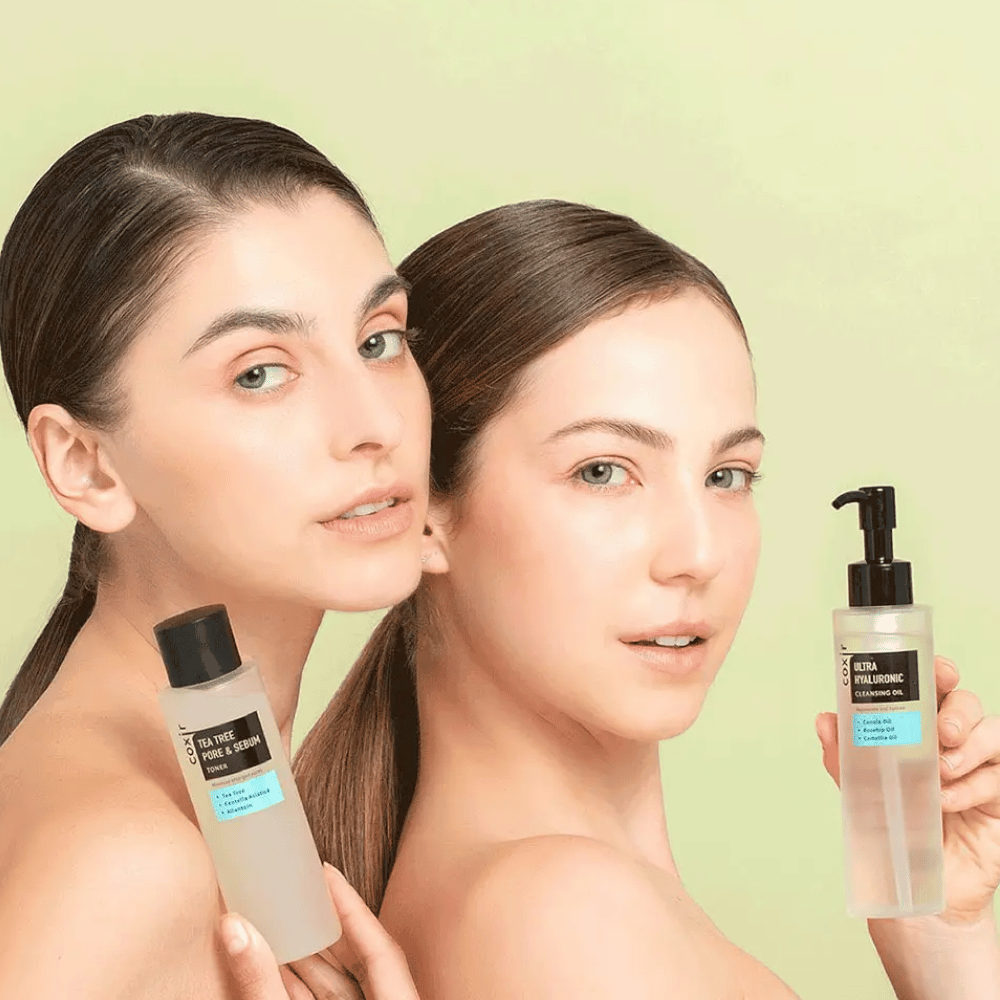 Zwei Frauen mit freien Schultern posieren mit Hautpflegeprodukten; die eine hält eine Flasche mit der Aufschrift "TEA TREE PORE & SEBUM", und die andere eine Pumpflasche mit der Aufschrift "ULTRA HYALURONIC CLEANSING OIL". Sie stehen vor einem hellgrünen Hintergrund.