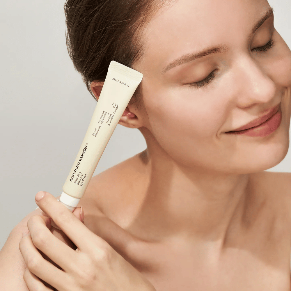 Eine lächelnde Person mit geschlossenen Augen hält eine Tube Haruharu Wonder Black Rice Bakuchiol Eye Cream an die Schläfe.