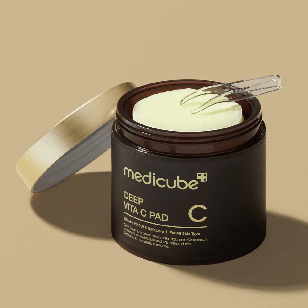 Eine Dose mit medicube Deep Vita C Pad. Die Dose ist dunkelbraun mit goldener Schrift und einem weißen, invertierten Etikett mit Vitamin-C-Informationen. Die Innenseite des Deckels ist golden, und eine durchsichtige Pinzette liegt auf den hellgelben Baumwollpads im Inneren. Der Hintergrund ist einfarbig beige.